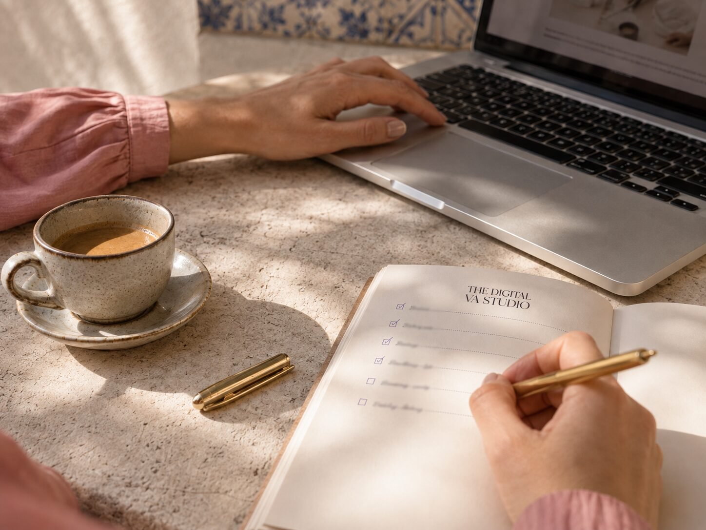Hands writing in a branded notebook beside a laptop and coffee in soft daylight